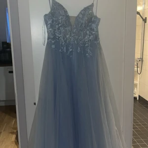 Ljusblå långklänning med spetsdetaljer - Magisk ljusblå långklänning med tunna axelband, djup v-ringning och vackra spetsapplikationer med blommönster upptill. Kjolen är i flera lager tunn tyll som ger en drömmig känsla. Snörning i ryggen för perfekt passform. Perfekt för bal eller student!