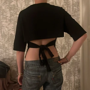 Svart croppad topp med knyt i ryggen NELLY - Säljer en svart croppad topp med halvlånga ärmar och öppen rygg som knyts baktill. Toppens stilrena design gör den lätt att matcha och den sitter snyggt figurnära. Perfekt för dig som gillar trendiga och unika detaljer.
