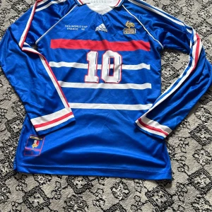 Frankrike Zidane VM 1998 långärmad tröja - Legendarsik blå långärmad fotbollströja från Adidas, Frankrikes landslag VM 1998 med Zidane 10 på ryggen. Klassiska vita och röda ränder, broderad tupp och matchdetaljer på bröstet. Snygg krage med röd, vit och blå kant. Perfekt för samlare eller fans.