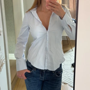 Ljusblå skjorta från Zara - Klassisk ljusblå skjorta från Zara med lång ärm, figursydd passform och knappar framtill. Skjortan har breda manschetter och en stilren krage. Perfekt att styla med jeans eller kjol för en clean look.
