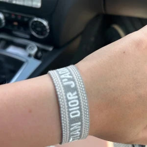 Dior armband - Snyggt vävt armband från Christian Dior i ljusgrått med vit text 'J'ADIOR'. Armbandet har justerbar knytning med tofsar och bred design som ger en trendig look. Perfekt accessoar för att lyfta din outfit med en exklusiv touch.