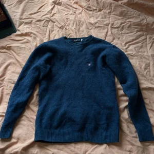 Mörkblå ulltröja från GANT - Snygg mörkblå stickad tröja från GANT med rund halsringning och broderad logga på bröstet. Tröjan är i mjuk ull och har ribbade muddar vid ärmslut och nederkant. Perfekt för dig som gillar klassisk och stilren look. Väldigt liten i storlek.