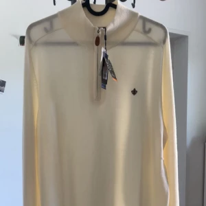 Morris Tröj - Snygg ljusbeige långärmad tröja från Morris i 100% merinoull. Tröjan har half zip med läderdetalj, hög krage och en broderad logga på bröstet. Perfekt för dig som gillar stilrena och klassiska plagg med premiumkänsla.