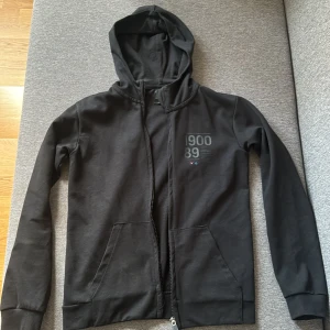 Svart hoodie med dragkedja från SOC - Svart hoodie från SOC med tryck '1900 89' på bröstet. Tröjan har huva, långa ärmar, två fickor fram och hel dragkedja. Tillverkad i mjuk bomullsmix, perfekt för chill eller träning. Enkel och stilren design som funkar till det mesta.
