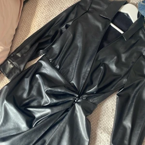 Svart kort omlottklänning i skinn med en slits - Snygg svart omlottklänning från Zara i skinn med lång ärm och markerad midja med twist-detalj framtill. Klänningen har djup v-ringning och klassisk krage.