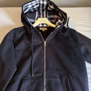 Svart hoodiejacka från Burberry - Svart hoodi från Burberry med klassiskt rutigt foder i huvan. Jackan har dragkedja framtill, snörning i huvan och två fickor. Ikonisk Burberry-logga på bröstet och detaljer i silver. Pris går alltid att diskutera