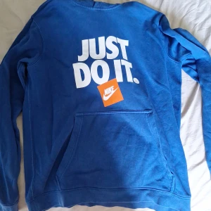 Blå Nike hoodie med tryck, stl M - Blå hoodie från Nike med stor vit 'JUST DO IT.'-text och orange Nike-logga på bröstet. Klassisk känguruficka framtill och huva. Skön och sportig stil, perfekt för chill eller träning. Tillverkad i mjuk bomullsblandning.