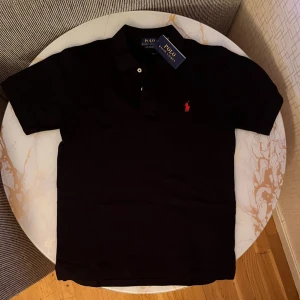 Svart pikétröja från Polo Ralph Lauren - Klassisk svart pikétröja från Polo Ralph Lauren med röd broderad logga på bröstet. Tröjan har korta ärmar, krage och knappar framtill. Perfekt för en stilren och sportig look.