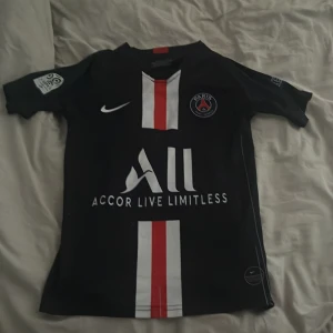 PSG Mbappé matchtröja Nike barn 140 - Paris Saint-Germain fotbollströja med Mbappé 7 på ryggen. Svart med vita och röda vertikala ränder, officiella klubbmärket och sponsortryck. Tillverkad i Dri-Fit material som andas. Korta ärmar och klassisk passform, perfekt för alla PSG-fans.
