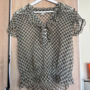 Blommig volangblus i transparent tyg - Supersöt blommig blus från Sfera i transparent, lätt material. Volangdetaljer vid krage, ärmslut och nederkant, samt knappar framtill. Mönstret är småblommigt i svart och gult på beige botten. 