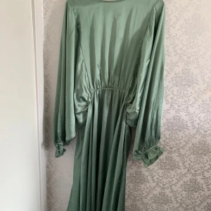 Grön långklänning från Bubbleroom XL - Snygg långklänning i ljusgrön satin från Bubbleroom. Klänningen har långa, vida ärmar med resår vid handlederna och markerad midja. Materialet är glansigt och ger en lyxig känsla. Perfekt för dig som vill sticka ut med en elegant och trendig look.