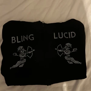 Svart hoodie med strassmotiv Bling Lucid - Svart hoodie med dragkedja och huva, dekorerad med strassmotiv av två amorfigurer och texten 'BLING' och 'LUCID' framtill. Hoddien har långa ärmar och är tillverkad i mjukt material, perfekt för en cool och avslappnad stil.