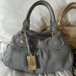 Grå Q Classic Baby Groove väska! - Så snygg grå blåaktig klassisk handväska i nyskick från Marc by Marc Jacobs i äkta läder med gulddetaljer och dubbla handtag. Väldigt snygg färg som passar till allt, den perfekta sommarväskan som rymmer mycket men är smidig och nätt. Lilla modellen Q Classic. Inga repor eller hål på väskans läder. Otroligt välbevarad vintage modell! Perfekt för att ge en stilren touch till din outfit. Skriv privat för fler bilder. Mått 34x18x12 (undersida). Kan hämtas hemma hos mig i Nacka eller skickas🤍