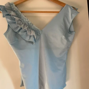 Ljusblå oneshoulder topp med volang - Supersöt ljusblå topp med oneshoulder-design och volangdetalj längs axeln. Perfekt för dig som vill sticka ut med en unik look 