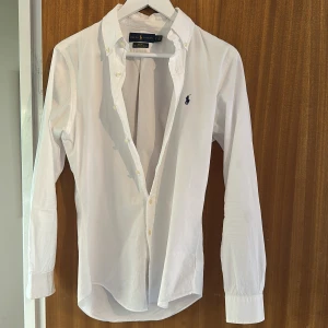 Vit slim fit skjorta från Ralph Lauren - Vit skjorta från Ralph Lauren i slim fit-modell, tillverkad i stretchig bomull. Klassisk design med knappar framtill, lång ärm och broderad marinblå logga på bröstet. Perfekt för en clean och stilren look.