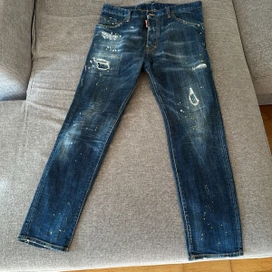 Dsquared2 blå jeans med slitningar - Snygga blå jeans från Dsquared2 med coola slitningar, färgstänk och distressed detaljer. Modellen är smal och har klassisk femficksdesign. Jeansen är tillverkade i bomull och har en modern streetkänsla med unika detaljer.
