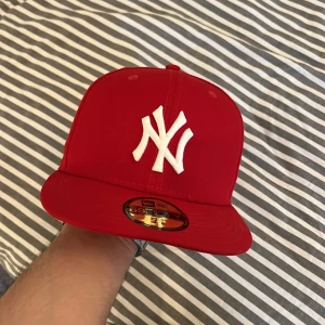 Röd New Era Yankees keps 59FIFTY - Snygg röd New Era 59FIFTY keps med klassiskt New York Yankees broderi i vitt framtill. Kepsen har rak skärm, broderade ventilationshål och MLB-logga bak. Tillverkad i polyester för en clean och sportig look.