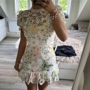 Blommig spetsklänning med volang - Superfin kort klänning i vit spets med blommigt mönster i pastellfärger som rosa, gul och grön. Puffiga korta ärmar och volang nertill ger en romantisk vibe. Perfekt för sommarens alla tillfällen! Öppen för bud 