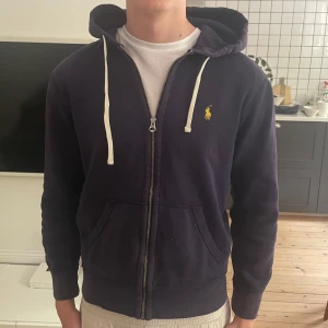 Mörkblå hoodie från Polo Ralph Lauren - Mörkblå hoodie från Polo Ralph Lauren med dragkedja, vit snörning i huvan och klassisk gul broderad logga på bröstet. Tröjan har känguruficka och är tillverkad i mjuk bomull. Perfekt för chill dagar och snygg till jeans.