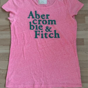 Rosa t-shirt från Abercrombie & Fitch - Snygg rosa t-shirt från Abercrombie & Fitch med stort grönt tryck på bröstet. Klassisk rund halsringning och korta ärmar. Perfekt för dig som gillar en enkel men ändå trendig look. T-shirten är i mjuk bomull och har en lätt melerad effekt. Hade på mig en gång. Köpt i USA. Storlek M, men är stort i storleken och passar L också.