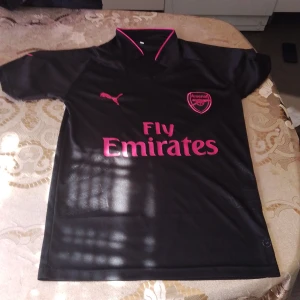 Arsenal svart fotbollströja Puma - Svart Arsenal fotbollströja från Puma med rosa detaljer och loggor. Tröjan har korta ärmar, V-ringning med rosa kant och tryckta sponsor- och klubbmärken. Tillverkad i lätt och ventilerande material, perfekt för match eller träning.