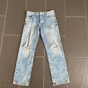 Blå slitna raka jeans med hål - Primark - Säljer ett par ljusblå jeans med raka ben och slitna detaljer, inklusive hål på båda knäna. Jeansen har klassisk femficksdesign och normal passform. Perfekta för en avslappnad och trendig look.