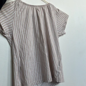 Randig blus i ljusrosa och vit - Söt blus från Etab med vertikala ljusrosa och vita ränder. Modellen har korta ärmar och en avslappnad passform med resår vid halsen. Perfekt för en chill och somrig look. Tillverkad i mjuk bomull som känns skön mot huden.