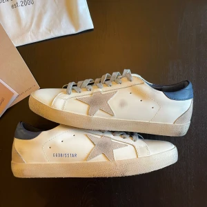 Golden Goose - Golden Goose Superstar sneakers i vitt skinn med grå mockadetaljer och stjärna på sidan. Grå snörning, blå hälkappa och beige sula ger en cool kontrast. Klassisk låg modell med rund tå och retrovibbar. Perfekta för dig som gillar streetstyle. Köpte dem några månader sedan och insåg att det var fel storlek. Tänkte kan ha dem tills de passar, men hände aldrig. I princip helt nya, bara testade på dem under tiden. 