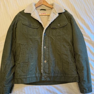 Grön manchesterjacka från Lee - Säljer en snygg grön manchesterjacka från Lee med mysigt teddyfoder. Jackan har knappar framtill och två bröstfickor med lock. Perfekt för kyliga dagar!