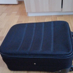 Svart resväska med hjul - Praktisk svart resväska med hjul och dragkedja. Perfekt för resor med sitt rymliga inre och smidiga hjul för enkel transport. Väskan har en hård yta och ett teleskophandtag för bekväm hantering.