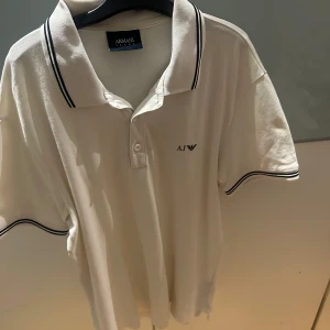 Vit pikétröja från Armani Jeans - Säljer en stilren vit pikétröja från Armani Jeans med klassisk krage och knappar. Tröjan har svarta detaljer på kragen och ärmsluten samt en liten logotyp på bröstet. Perfekt för en avslappnad men ändå elegant look.