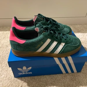 Adidas Gazelle - Snygga Adidas Gazelle sneakers i grön mocka med rosa detaljer. Perfekta för att ge din outfit en färgklick! De är storlek 38 & 2/3 och är helt oanvända. Skriv om ni har frågor💚💕