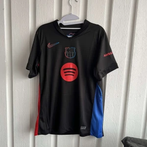 Svart fotbollströja från Nike - Säljer en svart fotbollströja från Nike med klubbmärke och sponsorlogga på framsidan. Tröjan har röda och blå detaljer på sidorna och korta ärmar. Perfekt för fotbollsfans som vill visa sitt stöd.