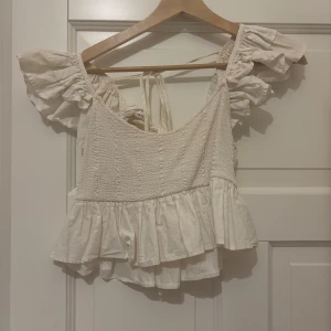 Vit blus från Zara - Charmig vit blus från Zara med volangdetaljer och korta ärmar. Toppen har en smockad design och knytband i ryggen för en justerbar passform. Perfekt för en somrig look.