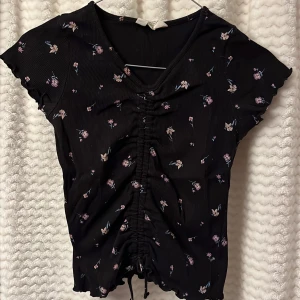 Svart blommig topp från H&M🖤 - Säljer en svart blommig topp från H&M med korta ärmar och en söt rynkad detalj framtill. Toppen har ett diskret blommönster i rosa och lila nyanser. 🖤🪻