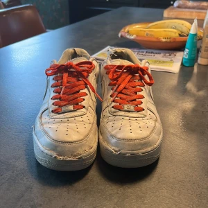 Nike Air Force Supreme - Säljer ett par vita Nike Air Force med röda snören och Supreme-detaljer. Skorna har en klassisk design med perforeringar på tån och en robust sula. Perfekta för dig som gillar en streetwear-look.
