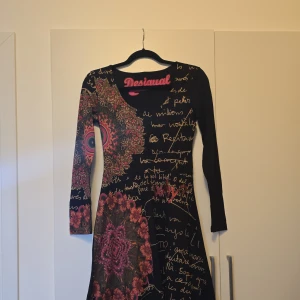 Svart klänning med mönster från Desigual - Snygg svart klänning från Desigual med unikt mönster i färgglada toner och textdetaljer. Klänningen har långa ärmar och en rund halsringning. Perfekt för dig som vill sticka ut med en kreativ stil.