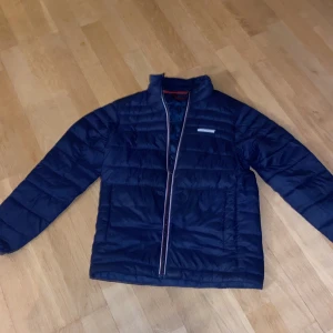 Mörkblå quiltad jacka från Jack & Jones - Säljer en snygg mörkblå quiltad jacka från Jack & Jones. Jackan har en dragkedja framtill med röd och vit kant, och är perfekt för kyligare dagar. Den har långa ärmar och en stilren design.