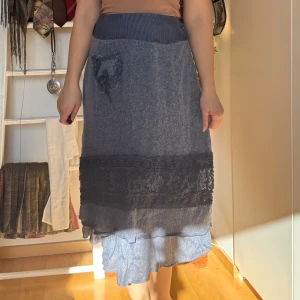 Blå stickad kjol med spetsdetaljer - Charmig blå stickad kjol med volang och spetsdetaljer. Kjolen har en unik design med en liten ficka framtill och en bred ribbad midja för extra komfort. Perfekt för en bohemisk look.