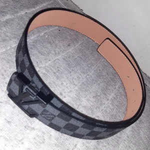 Rutig bälte med monogramspänne - Snyggt bälte med ett rutigt mönster och ett stilrent monogramspänne. Bältet har en klassisk design med en modern touch, perfekt för att lyfta vilken outfit som helst.
