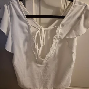 Vit blus med spetsdetaljer - Säljer en söt vit blus med spetsdetaljer och knytning i ryggen. Blusen har korta ärmar med volang och en v-ringad design. Perfekt för en somrig look!