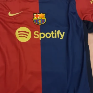 Blåröd fotbollströja från Nike - Säljer en blåröd fotbollströja från Nike med FC Barcelona-logga och Spotify-sponsor. Tröjan har korta ärmar och är tillverkad i Dri-FIT-material för optimal komfort. Perfekt för fans av laget! 💙❤️