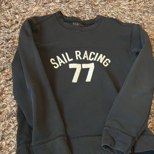 Svart tröja från Sail Racing - Säljer en svart tröja från Sail Racing med texten 'SAIL RACING 77' i vitt på framsidan. Tröjan har en klassisk design med långa ärmar och rund hals. Perfekt för en avslappnad stil.