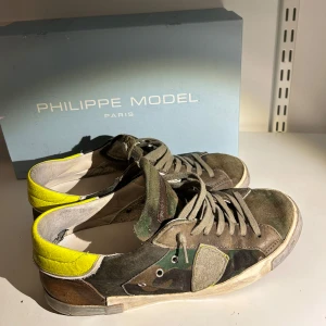 Camouflage sneakers från Philippe Model - Snygga sneakers från Philippe Model i en cool camouflage-design med inslag av gult och brunt. Skorna har snörning och är tillverkade i skinn och mocka. Perfekta för en trendig look. Tveka inte att höra av dig vid funderingar!