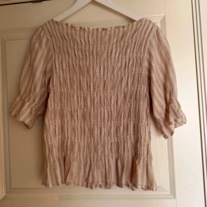 Abercrombie fitch peplum topp - Säljer en söt beige blus med ränder och trekvartsärmar. Blusen har en smockad design som ger en fin struktur och en lätt volangdetalj vid nederkanten. Perfekt för vår och sommar!