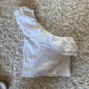 Vit oneshoulder topp med spetsdetaljer - Säljer en stilren vit oneshoulder topp med vacker spetsdetalj. Toppen har en ribbad struktur och en volang som ger en romantisk touch. Perfekt för sommarens alla tillfällen!