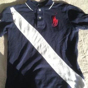 Blå pikétröja från Ralph Lauren - Säljer en snygg blå pikétröja från Ralph Lauren med vit diagonal rand och röd broderad logga. Tröjan har korta ärmar och knappar vid kragen. Perfekt för en stilren look!