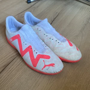 Vita och orange fotbollsskor från Puma - Säljer ett par vita fotbollsskor från Puma med orange detaljer. Skorna har en strumpliknande passform och är designade för inomhusbruk. Perfekta för snabba rörelser på planen.