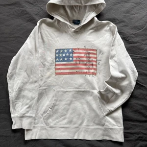 Vit hoodie från Polo Ralph Lauren - Säljer en vit hoodie från Polo Ralph Lauren med ett tryck av den amerikanska flaggan på framsidan. Perfekt för en avslappnad stil. Storleken är 14-16 år passar S 