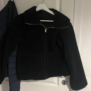 Svart teddyjacka med dragkedja - Mysig svart teddyjacka med dragkedja framtill. Perfekt för kyliga dagar med sin fluffiga och varma design. Jackan har långa ärmar och en enkel stil som passar till det mesta. Använt 2-3 gånger